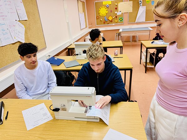 2 Schüler unter Anleitung einer Schülerin an der Nähmaschine